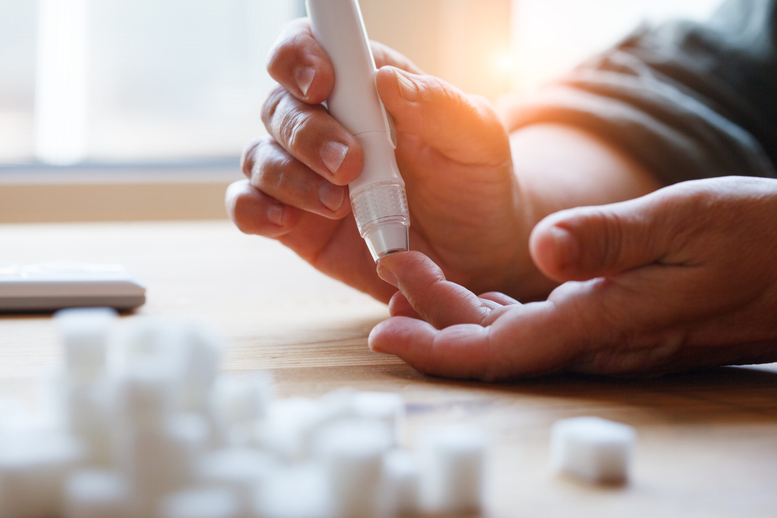 bftb21_03 senior woman hands using lancet on finger at home to check blood sugar level, glucometer and sugar cubes on wooden table close up, diabetes concept, elderly health care, sunny morning